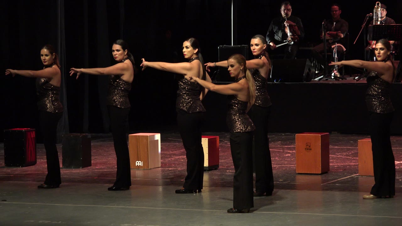 Women in dark sequined tops hold their arms out on a stage with musicians behind them.