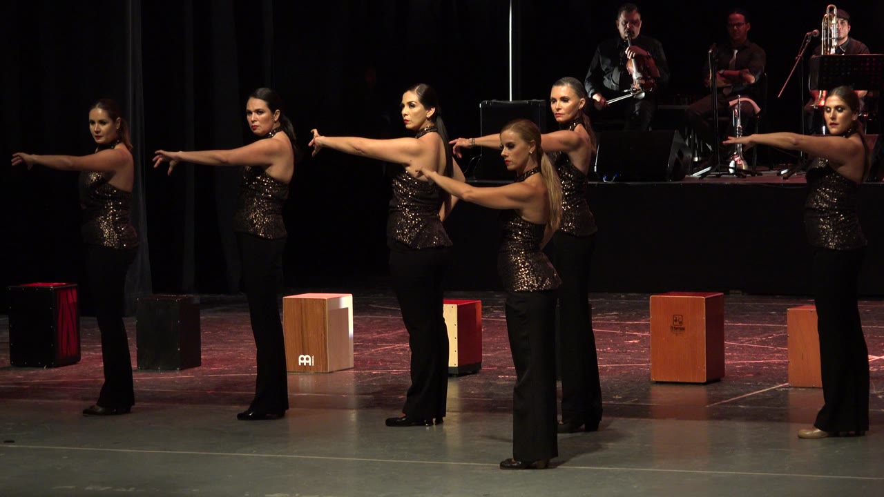 The dancers keep a side-facing arm extension on the Cabo San Lucas stage, with warm light catching their sequined tops and musicians behind them.