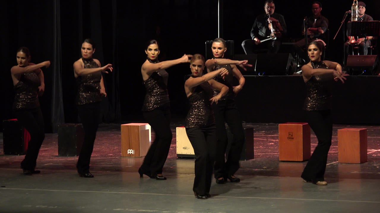 Women in matching dark outfits dance on a stage with musicians behind them.