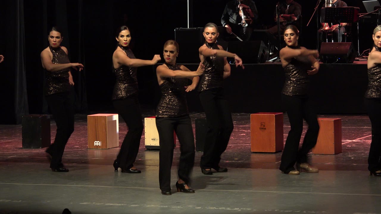 The dancers continue in formation on a Cabo San Lucas stage, with one performer centered and others extending arms in matching timing.
