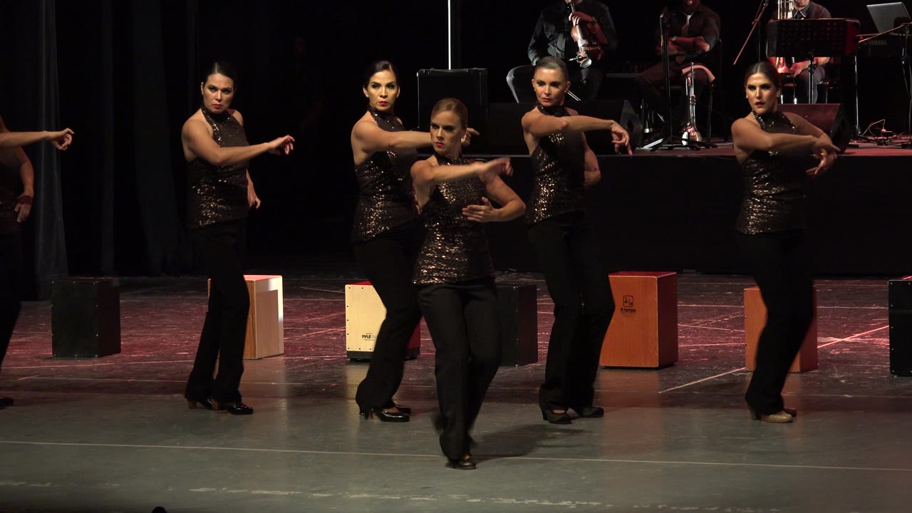 Women performing on a theater stage with musicians behind them.