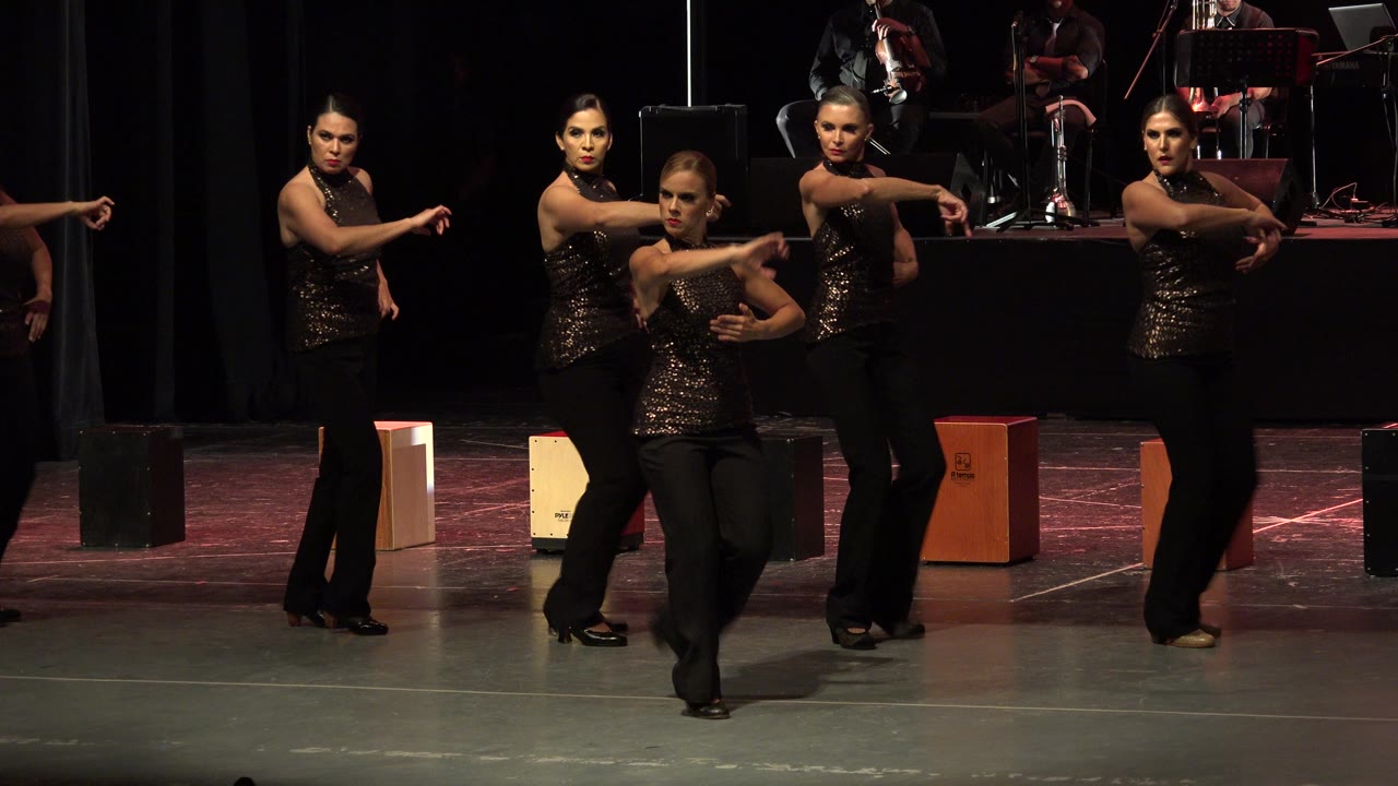 At 0:01, the dancers hold a synchronized pose with arms extended while musicians and percussion boxes remain visible at the rear of the Cabo San Lucas stage.