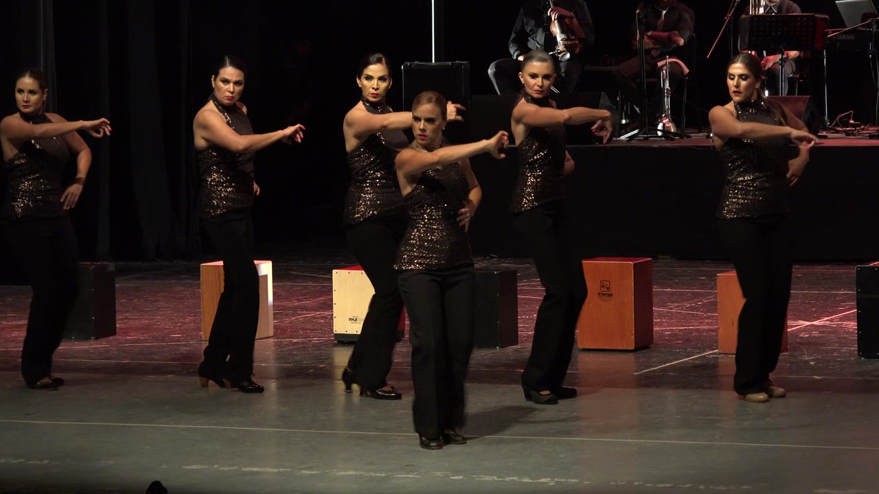 The dancers remain spread across the stage in Cabo San Lucas, Mexico, extending their arms forward while warm overhead light catches the sequined costumes.