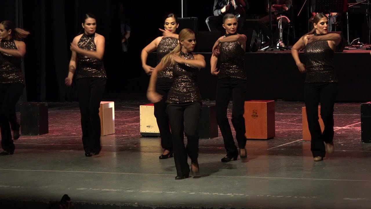 A lead adult dancer steps toward the camera during the Cabo San Lucas stage routine while other women in matching black outfits remain arranged behind her.