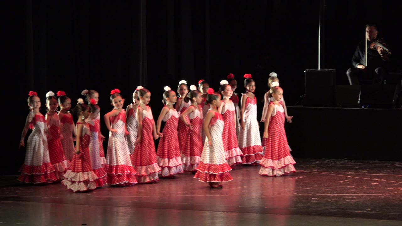 At the start of the Cabo San Lucas clip, girls in red and white ruffled dresses move across a dark stage under warm light with a seated musician in the background.