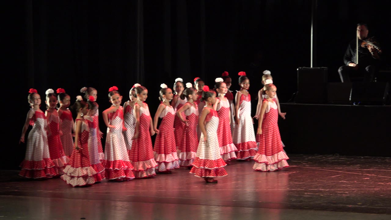 One second in, the dancers remain grouped across the midstage area, with several faces turned toward the front and the musician still visible above the stage line.