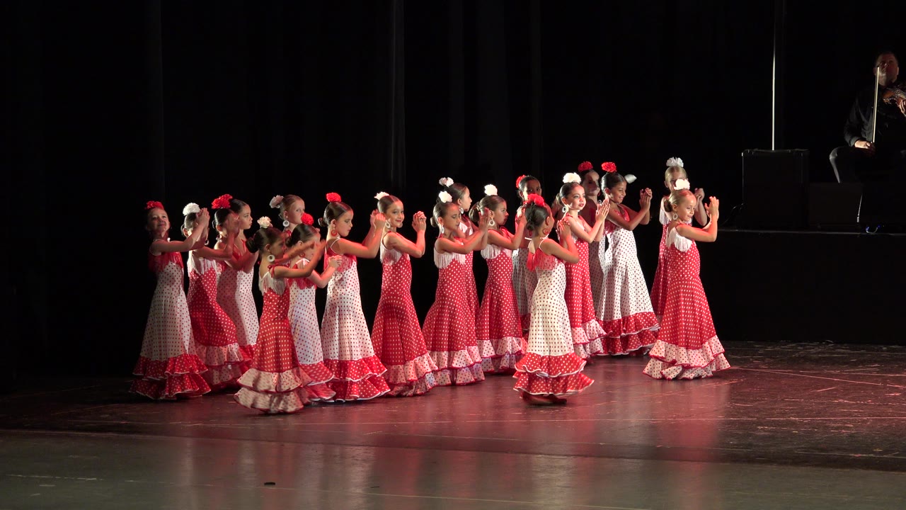 The dancers continue clapping across the theater stage in Cabo San Lucas, Mexico, with red bows and white hair accessories standing out under stage lights.