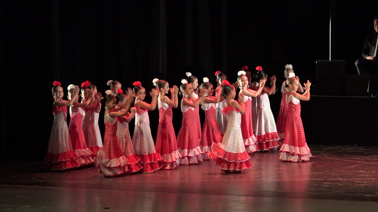 A group of children in long red and white patterned dresses clap together on a dark Cabo San Lucas stage.