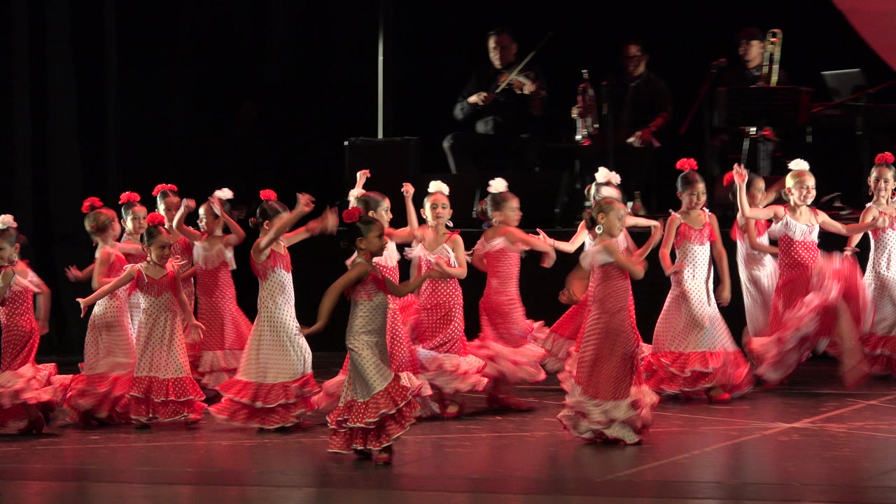 Young girls in pink dresses dancing on a stage with musicians behind them.