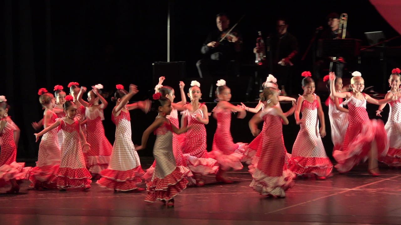 One second in, the dancers hold a loose line across the stage in Cabo San Lucas, with raised arms and pink costume ruffles catching red light.