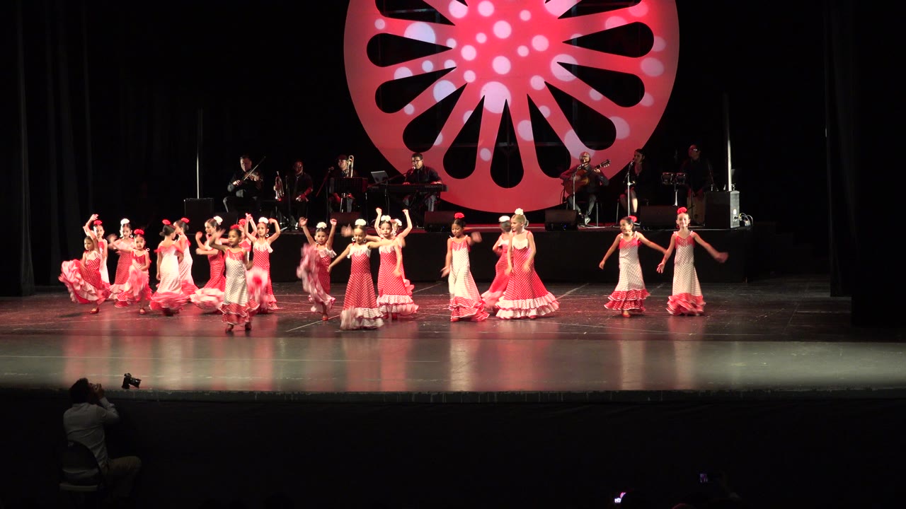 Wide view in Cabo San Lucas, Mexico, of children in pink, red, and white dance costumes spread across a stage beneath a large circular pink backdrop.