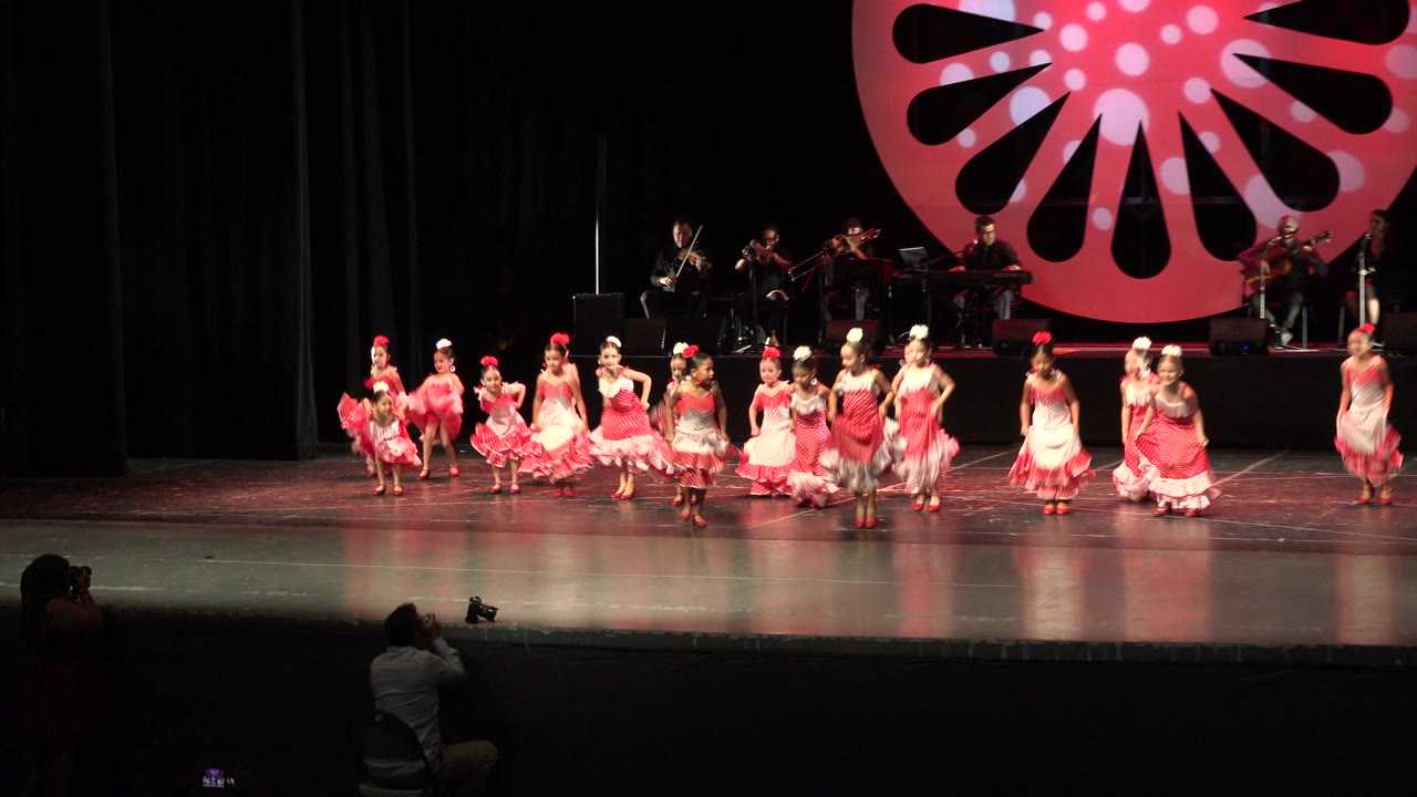 Girls in red and white dresses dance on a theater stage with musicians behind them.