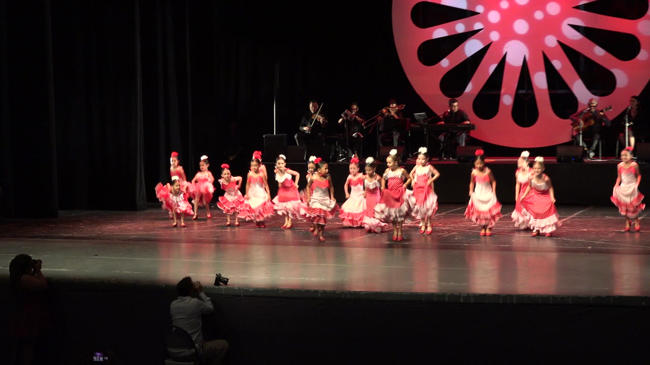 In Cabo San Lucas, Mexico, the dancers spread across the stage while the musicians remain on the riser behind them and a red decorative backdrop fills the upper frame.