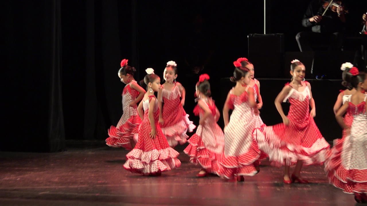 In Cabo San Lucas, Mexico, several young girls in red and white ruffled flamenco-style dresses move across a dark stage while a seated musician is partly visible above them.