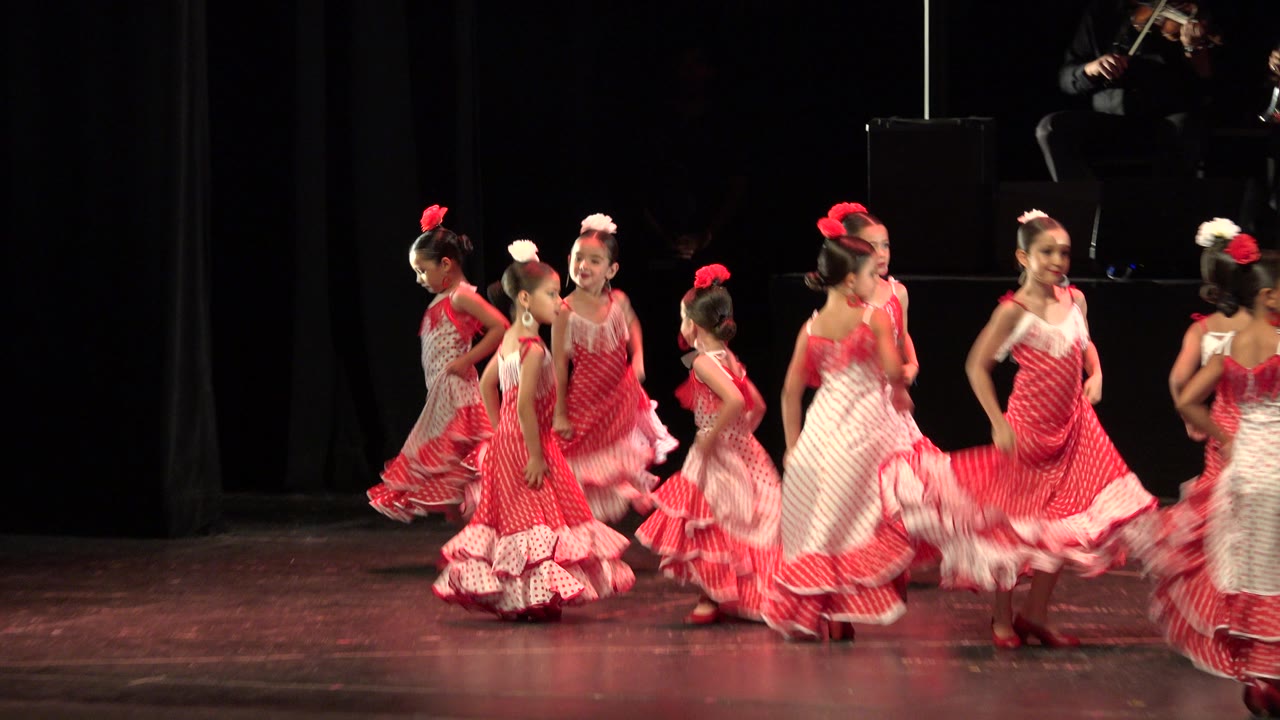 In Cabo San Lucas, Mexico, the children cluster near center stage in red and white dresses as one girl's skirt lifts during a turn and the black curtain remains behind them.