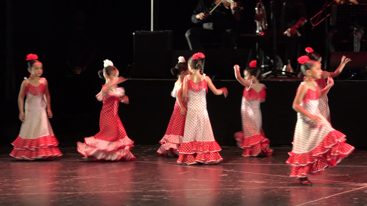 Young girls in red and white dresses dance on a dark stage.