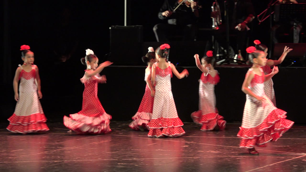 The dancers cross paths in Cabo San Lucas, Mexico, with one child turning in the foreground and others grouped behind under red stage light.