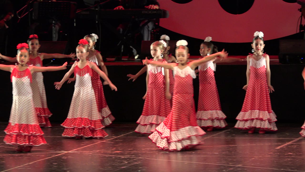 Young girls in red and white ruffled dresses perform on a stage in Cabo San Lucas, Mexico, with several arms extended and musicians dimly visible behind them.