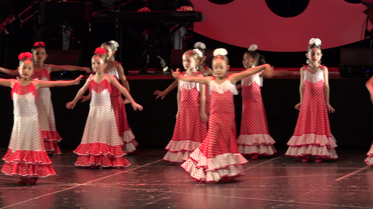 Several girls in coordinated red and white dresses hold out their arms during a stage dance in Cabo San Lucas, Mexico, with a dark band area behind them.
