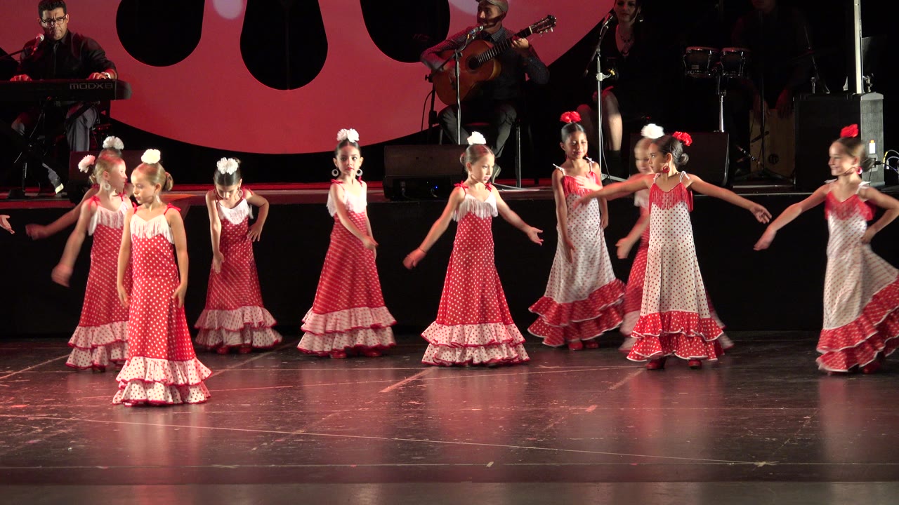 Young girls in red and white ruffled dresses stand and dance on a stage.