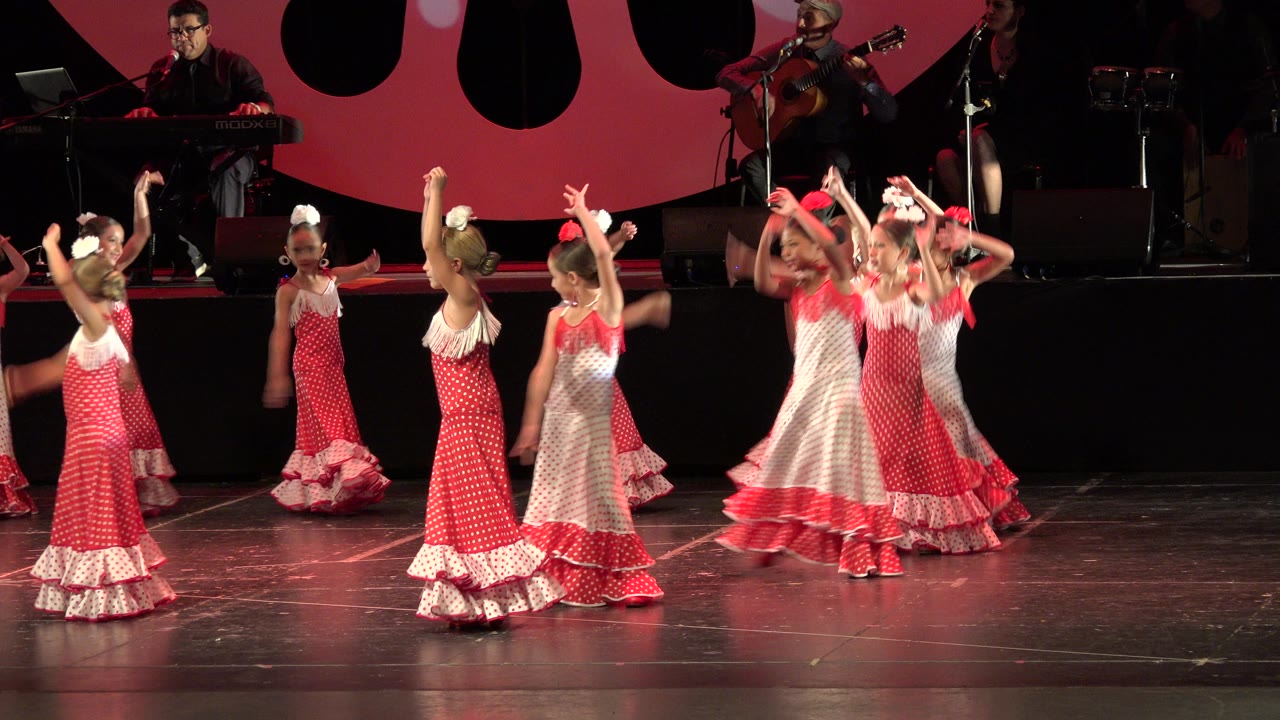 Young girls in red and white ruffled dresses dance in a loose group on a stage in Cabo San Lucas, Mexico, with musicians, microphones, and red backdrop shapes behind them.