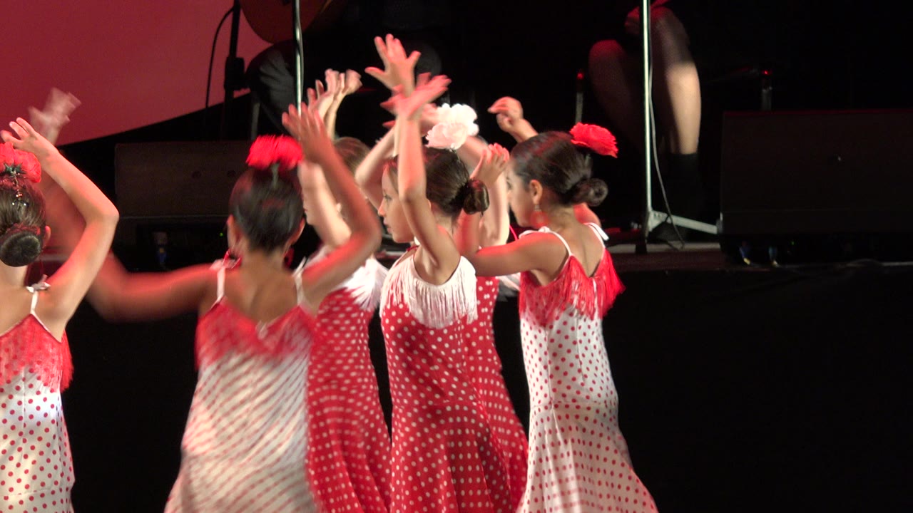 The group of girls remains close together with arms lifted, red and white dresses catching warm stage light in Cabo San Lucas, Mexico.