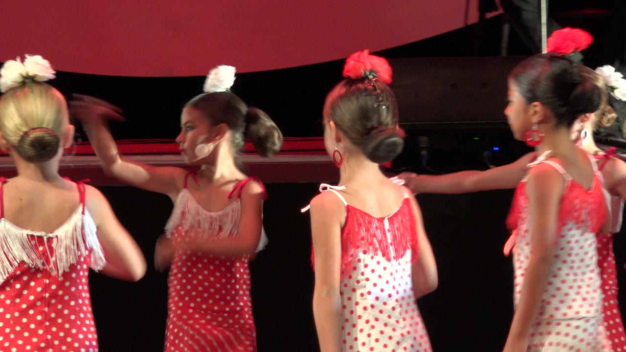 Several young dancers in matching red and white dresses turn across the stage in Cabo San Lucas, Mexico, with one blonde girl seen from behind and others moving past her.