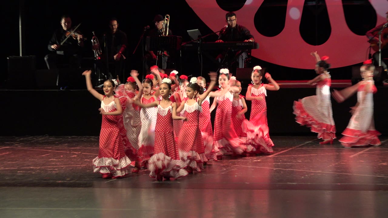 Children in red and white dresses dance on a stage with musicians behind them.