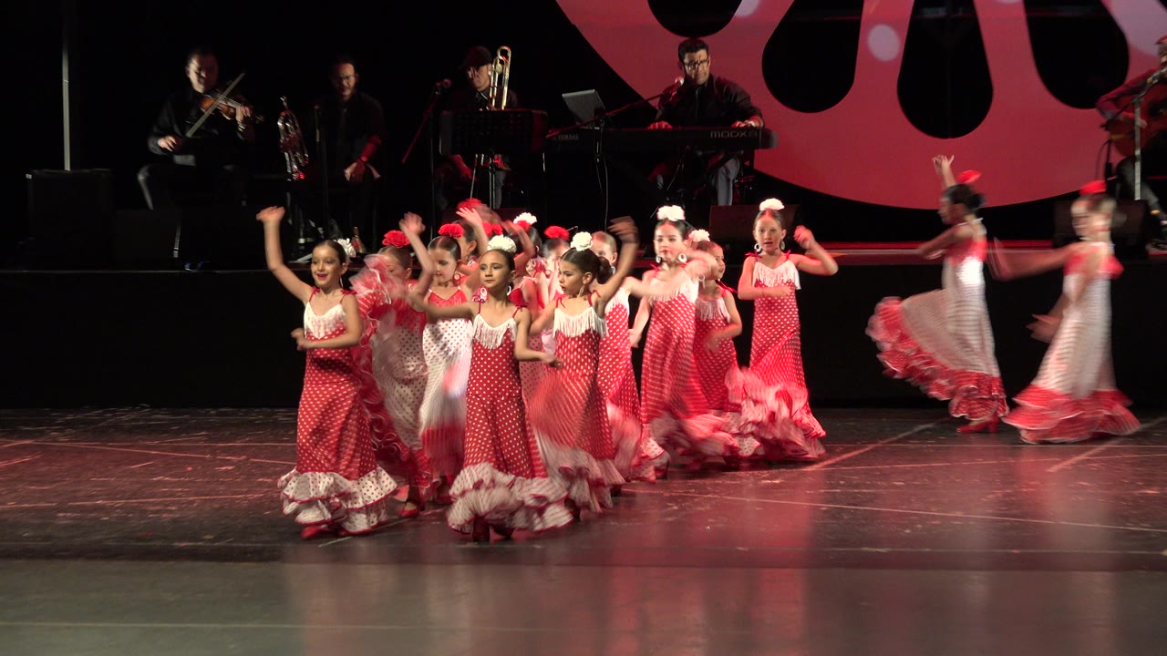 The dance line shifts across the stage in Cabo San Lucas, Mexico, with children raising their arms and a blurred dancer crossing near the right edge.