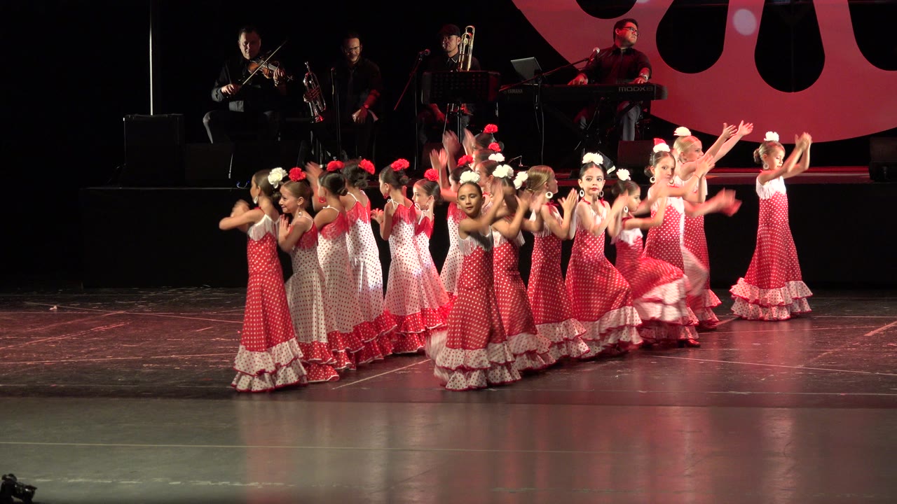 Girls in red and white dresses dance on a stage with musicians behind them.
