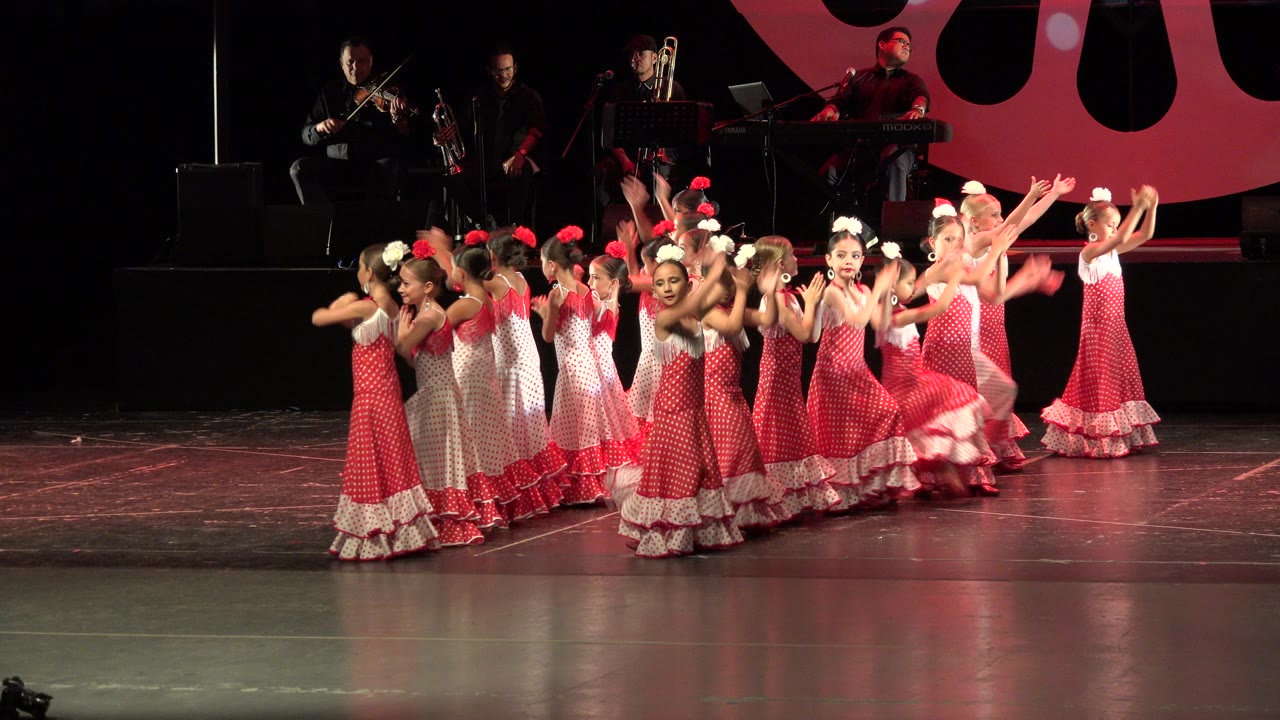 At 0:01, the dancers hold their arms up in coordinated motion on the Cabo San Lucas stage, with red skirts and white ruffles catching the warm performance light.