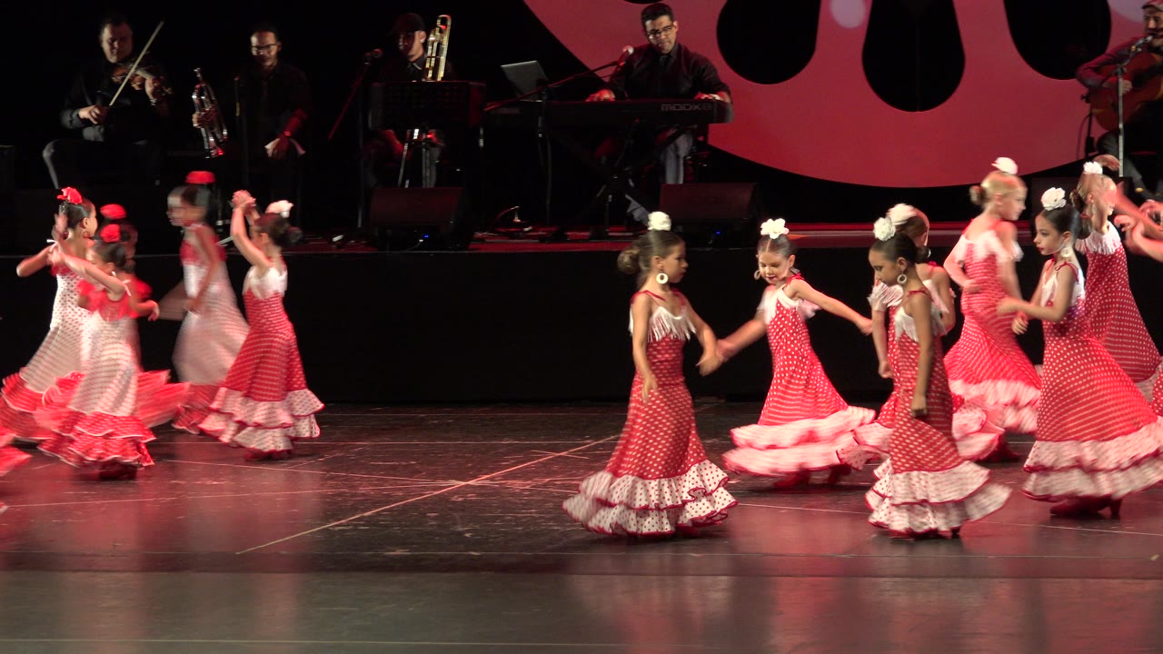 Girls in red and white dresses dance on a theater stage with musicians behind them.