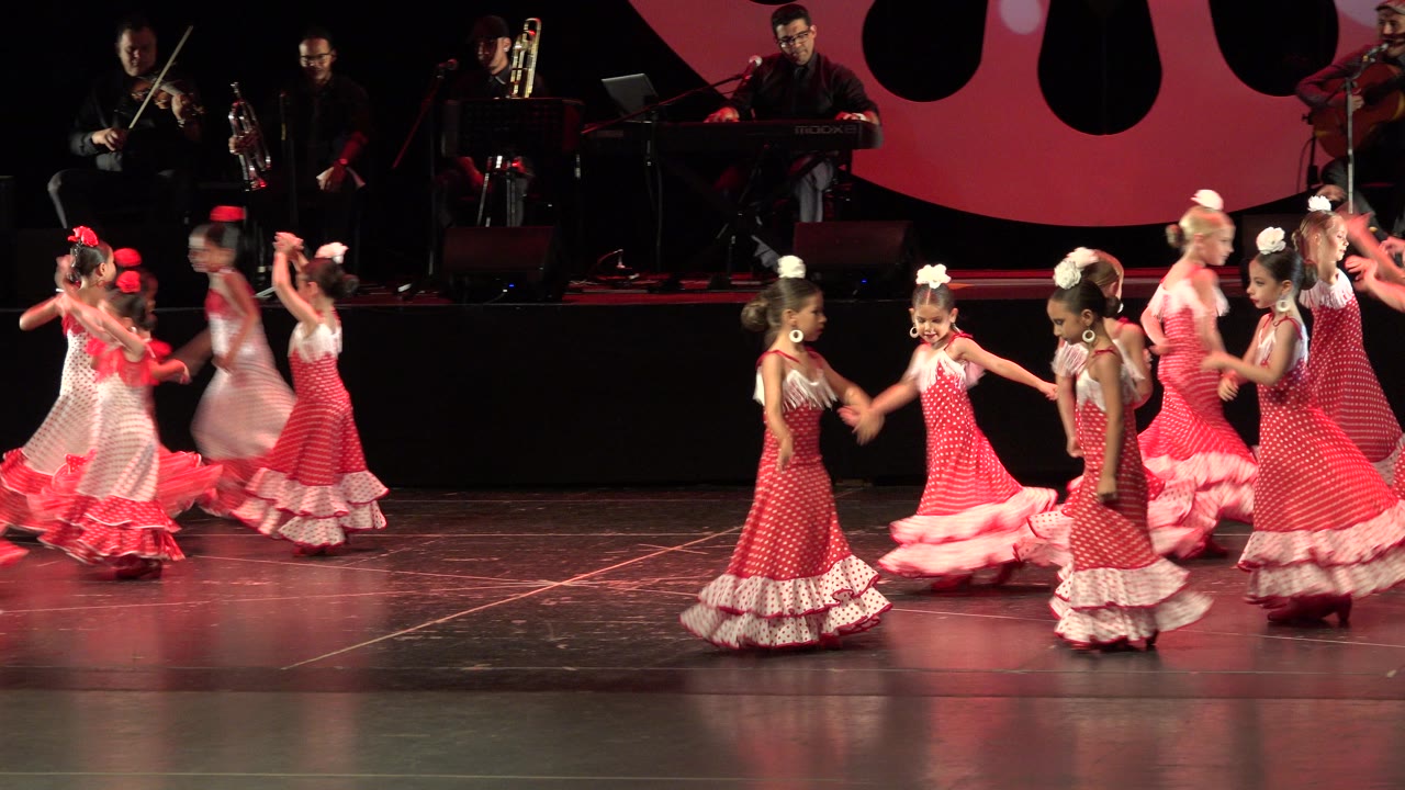 At 0:01 in Cabo San Lucas, Mexico, several girls in matching dresses face each other in pairs as other dancers move behind them on the lit stage.