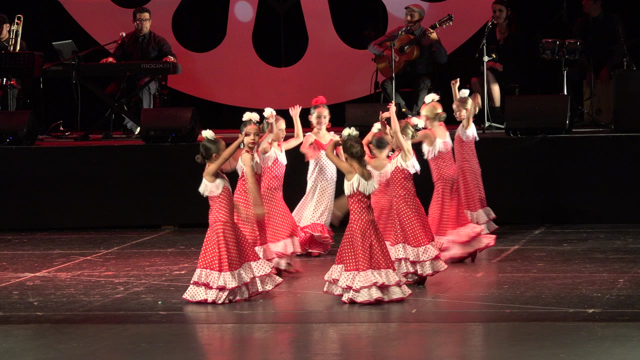 In Cabo San Lucas, Mexico, young girls in red and white ruffled dresses gather mid-dance on a dark stage while a keyboard player and guitarist are visible behind them.