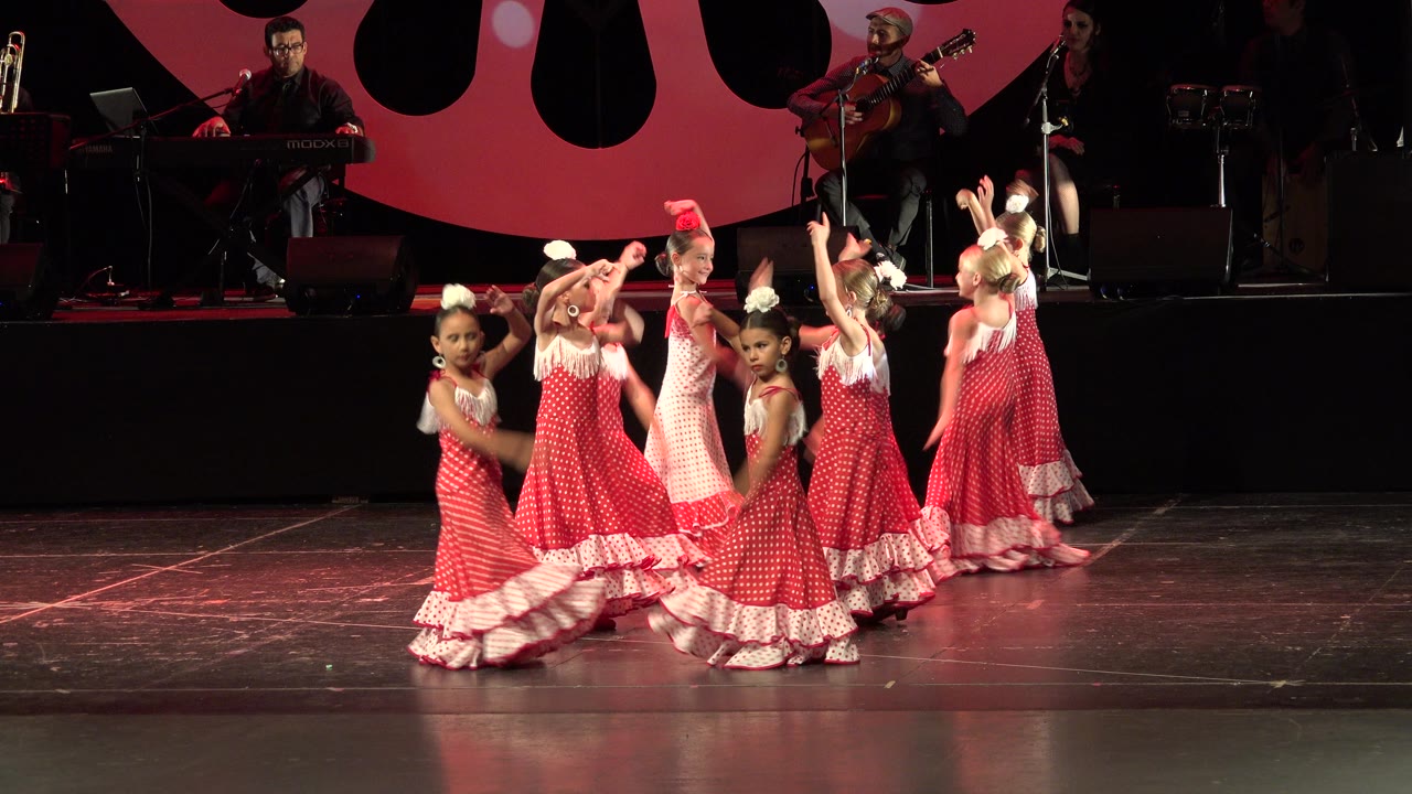 Girls in matching red and white dresses turn across a lit stage.