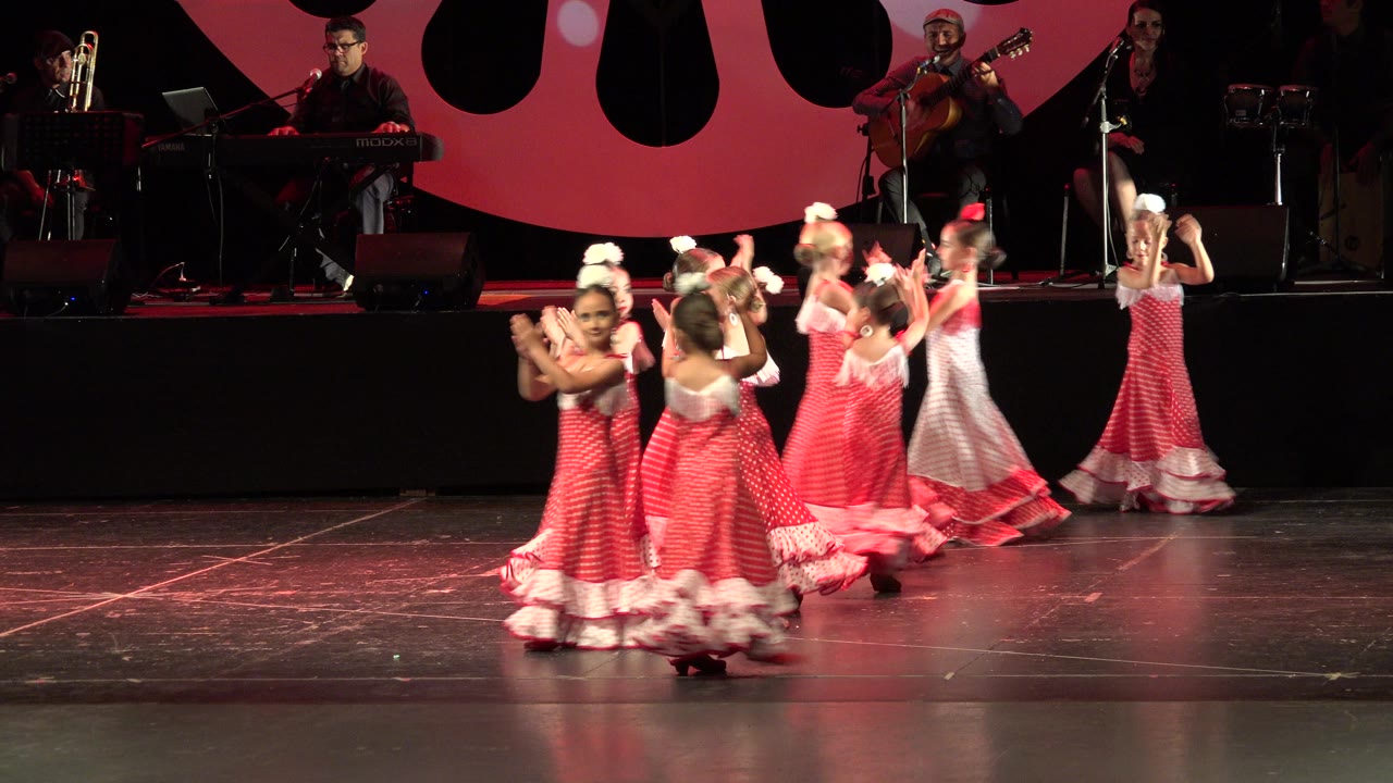 Children in red folk dresses dance on a stage with musicians behind them.