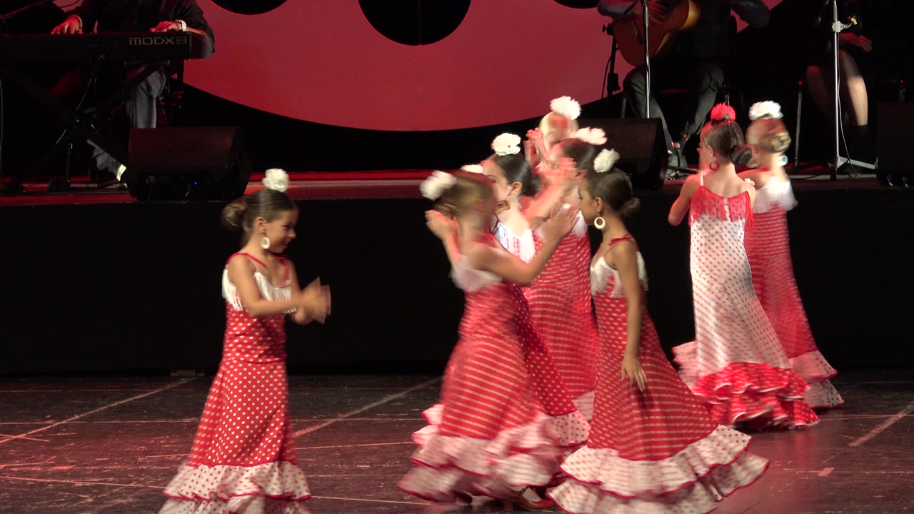 Girls in coordinated red and white dresses clap and cross paths on the dark stage during the Cabo San Lucas performance.