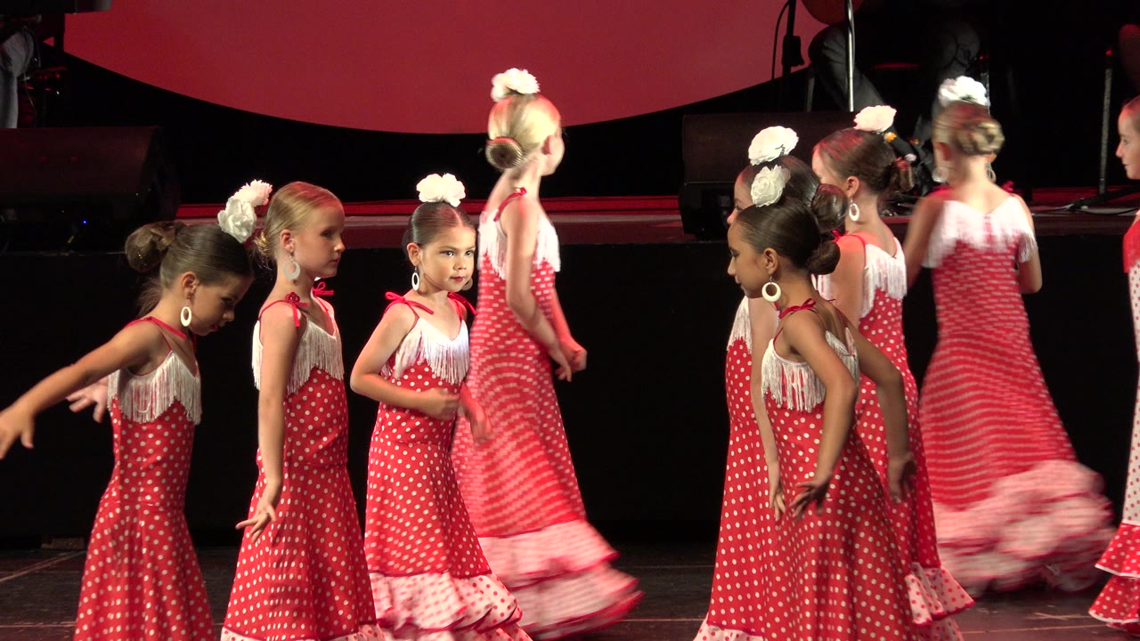 Young girls in matching red polka-dot dresses gather and move across a dark stage during a 2019 Cabo San Lucas dance performance.