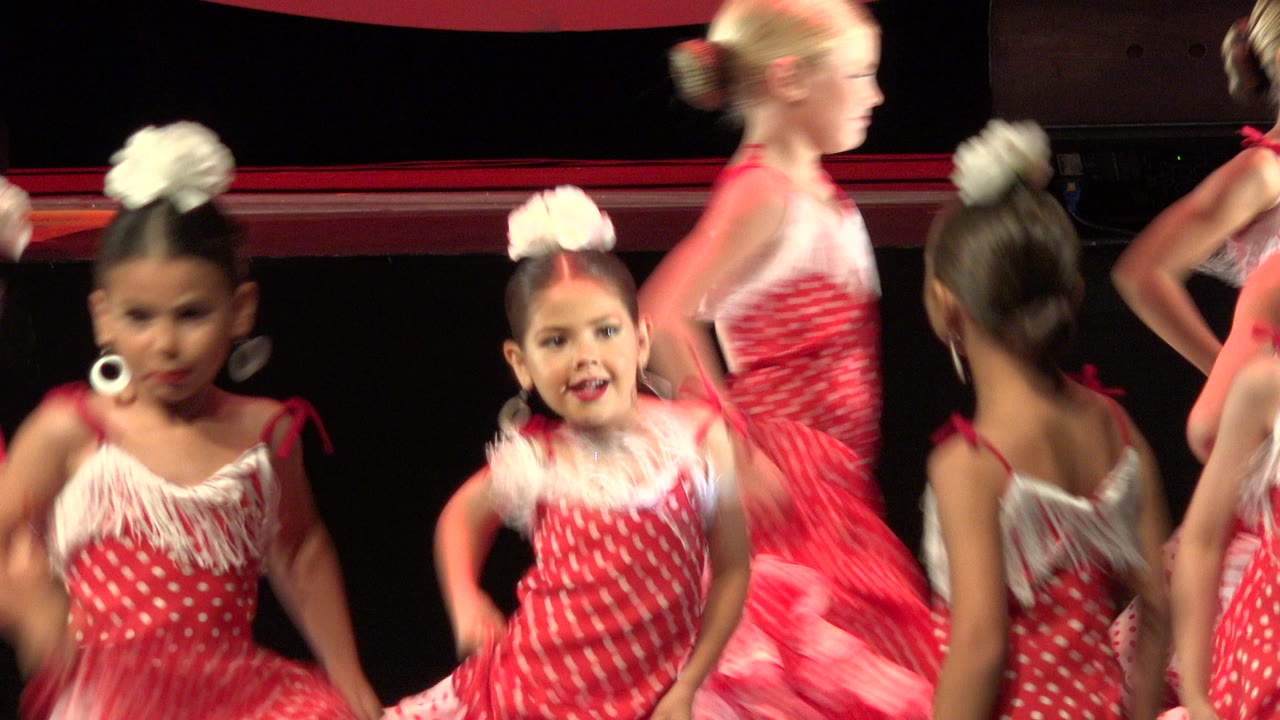 Several young dancers in red and white costumes cross the stage in Cabo San Lucas, Mexico, with a blonde child passing through the center and another child facing camera.