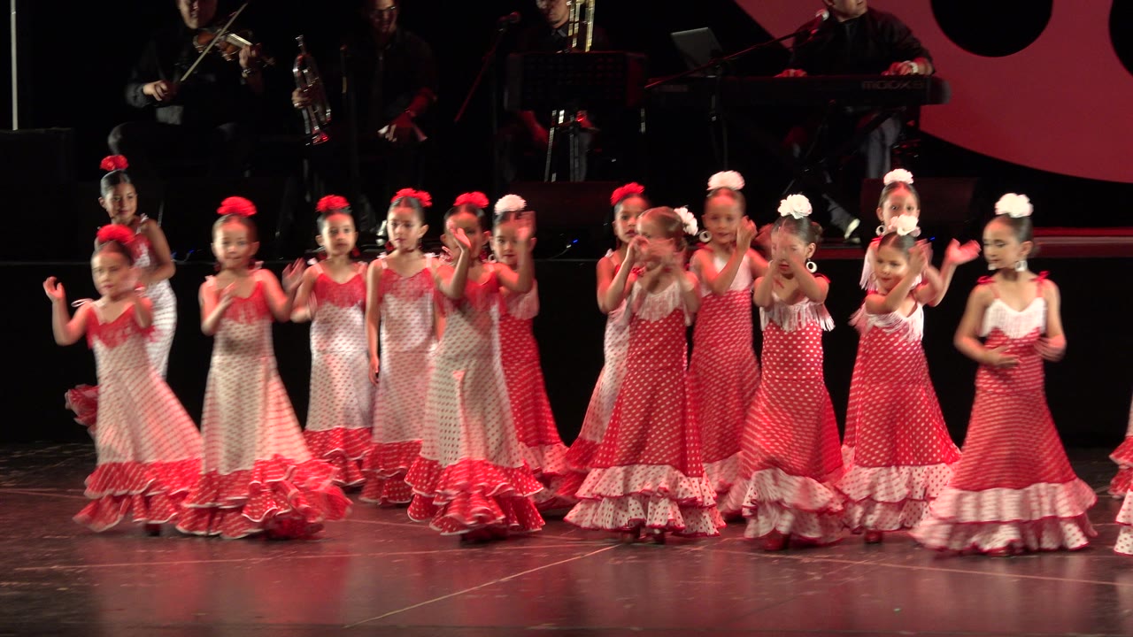 Young girls in red and white polka-dot recital dresses clap together on a dark stage in Cabo San Lucas, Mexico, with musicians dimly visible behind them.