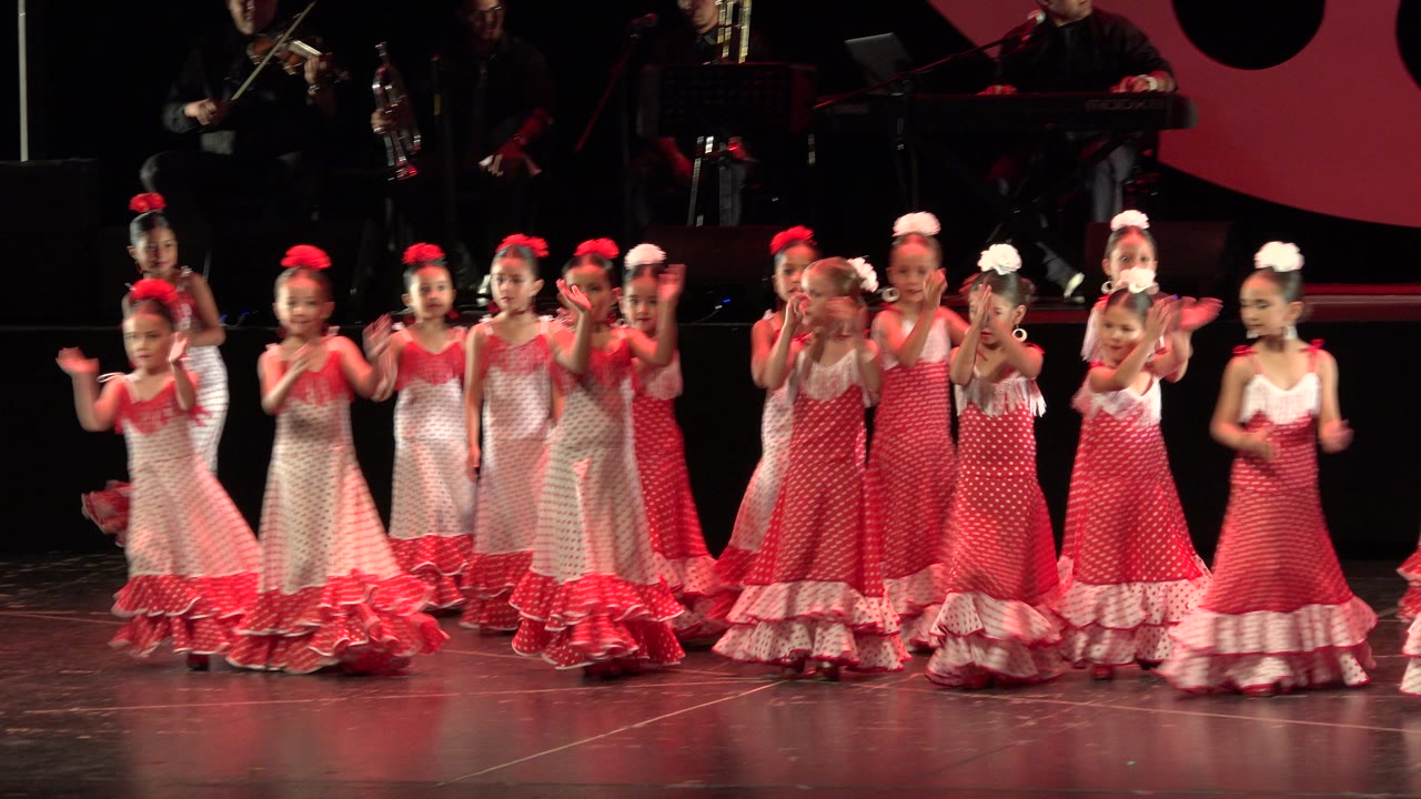 Children in matching red and white dresses continue clapping in a line on the Cabo San Lucas stage, with red lighting and shadowed musicians in the background.