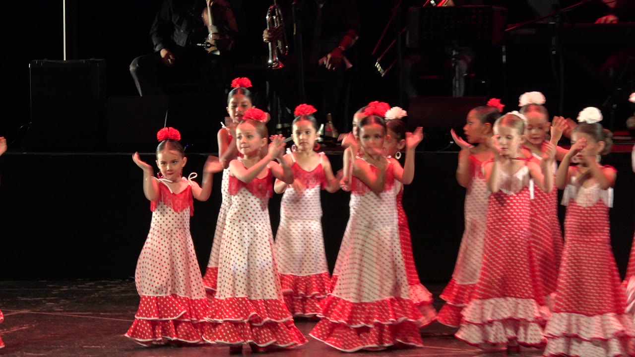 Children in red and white stage dresses clap on a dark theater stage.