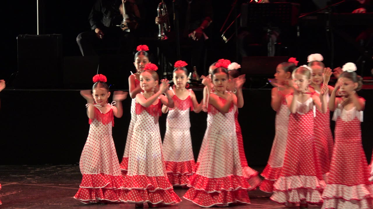 Young performers in matching red and white costumes clap near the front of the stage, with partially visible band musicians behind them in Cabo San Lucas.