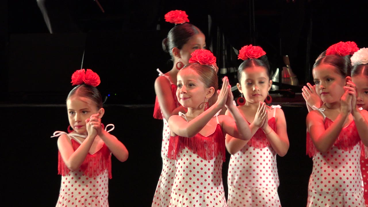Young girls in red and white dance costumes clap on a dark stage.