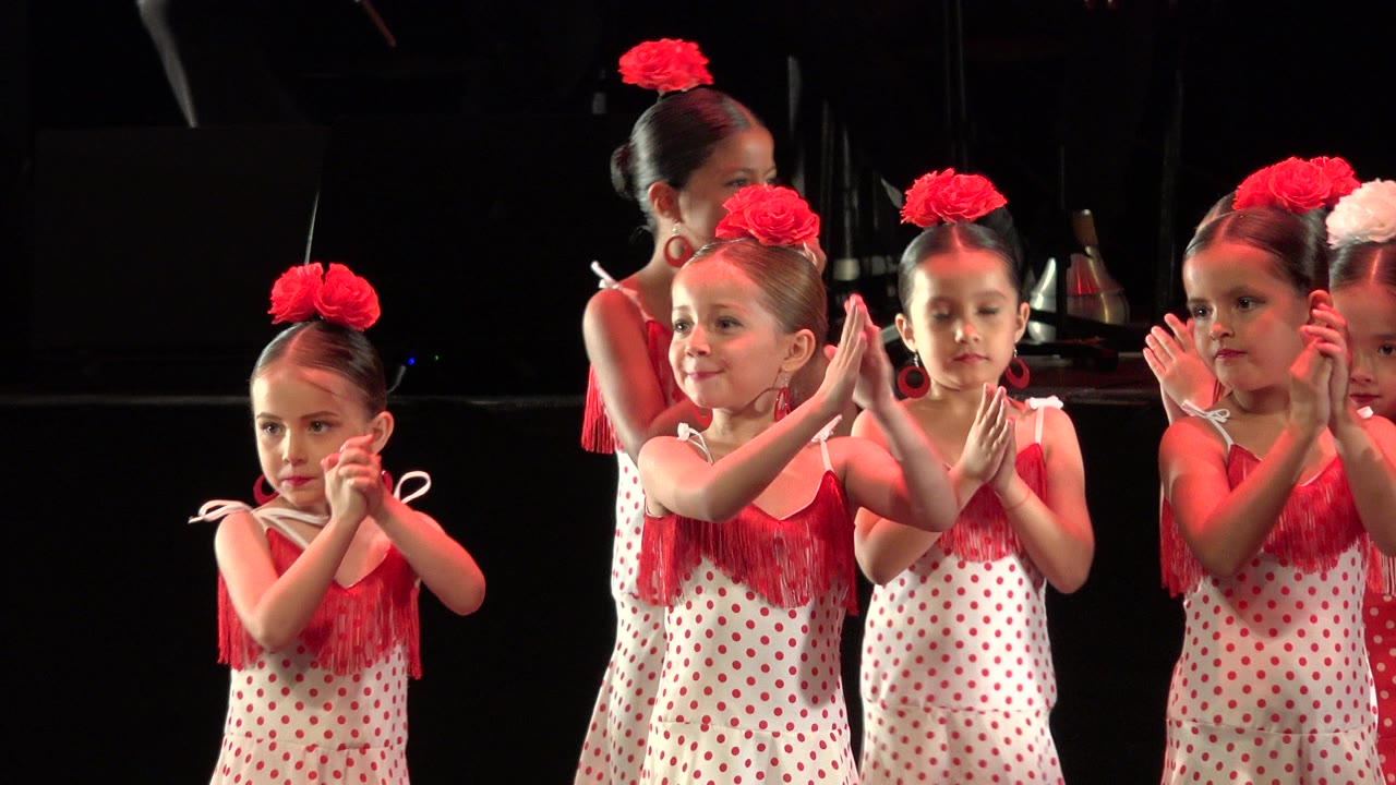Several young performers in red-and-white dance dresses clap in place on a dark stage in Cabo San Lucas, Mexico, with one older girl partly visible behind them.