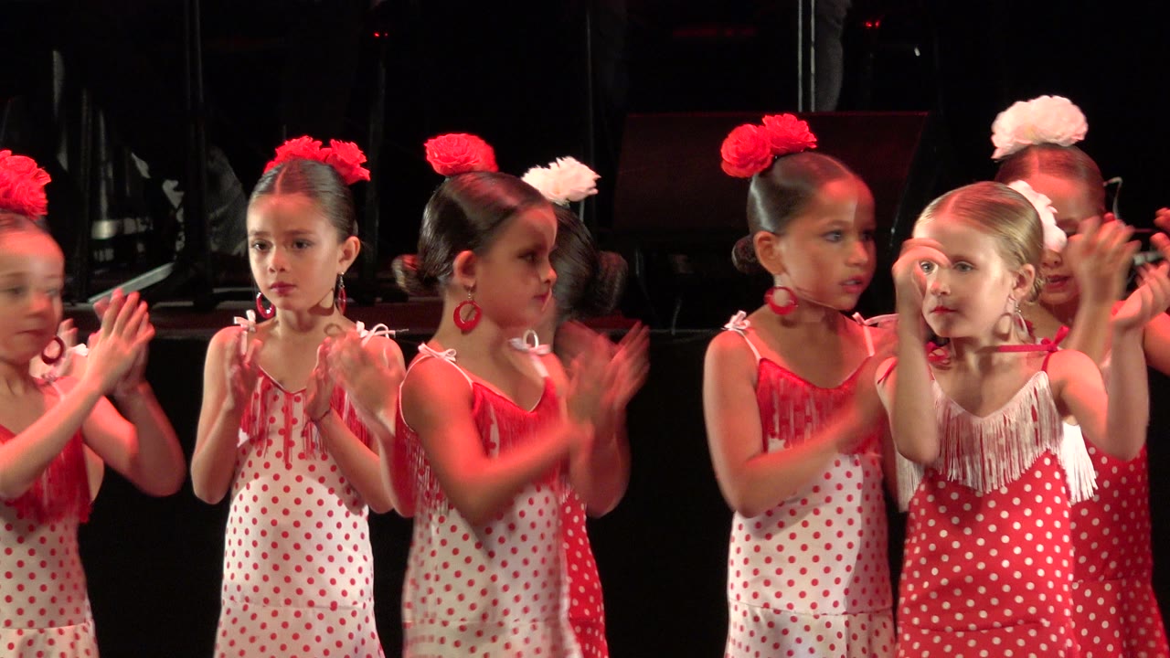 Young girls in red and white dresses clapping on a dark stage.