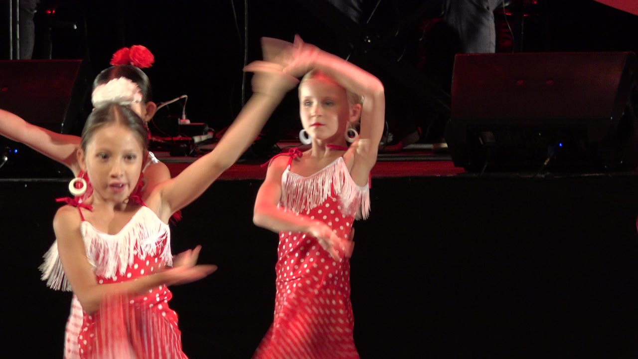 Two young girls in red polka-dot dance dresses raise their arms on a dark stage.