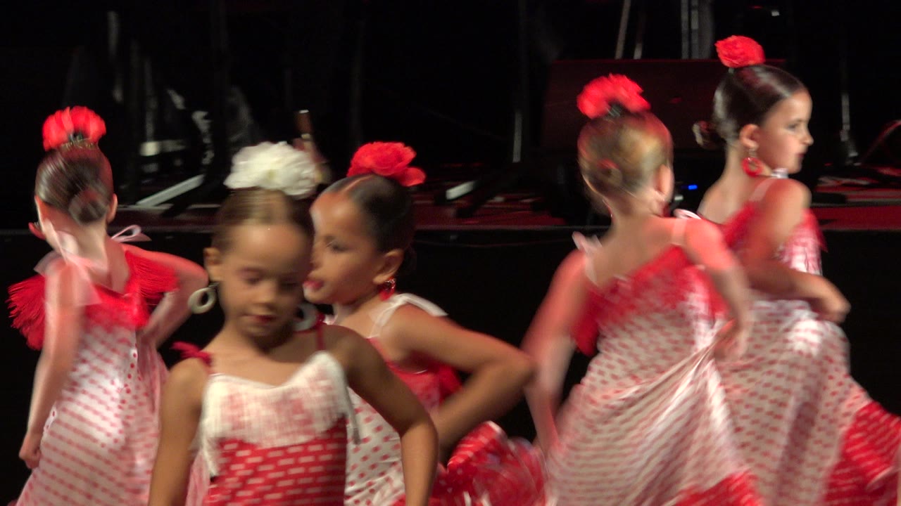Girls in red and white dresses dance on a dark stage.