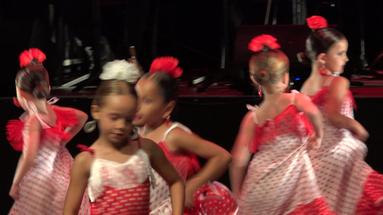 At 0:01, several girls in red and white dance costumes turn and pass across the stage, with red hair accessories catching the warm light.