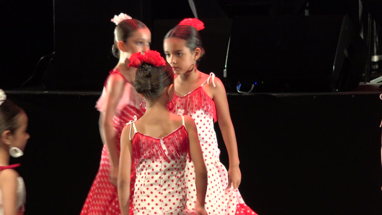 Girls in red and white polka-dot dresses standing on a dark stage.